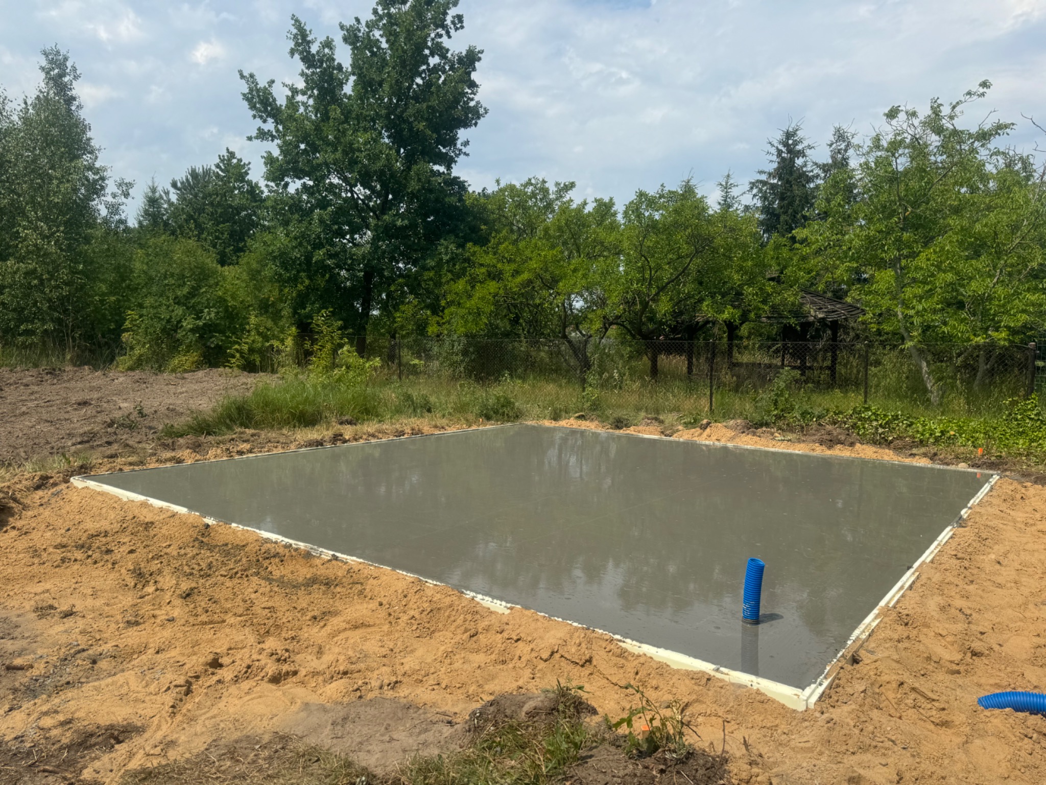 Wylany betonowy fundament płyty na działce z widocznymi niebieskimi rurami wentylacyjnymi i otaczającą zielenią.