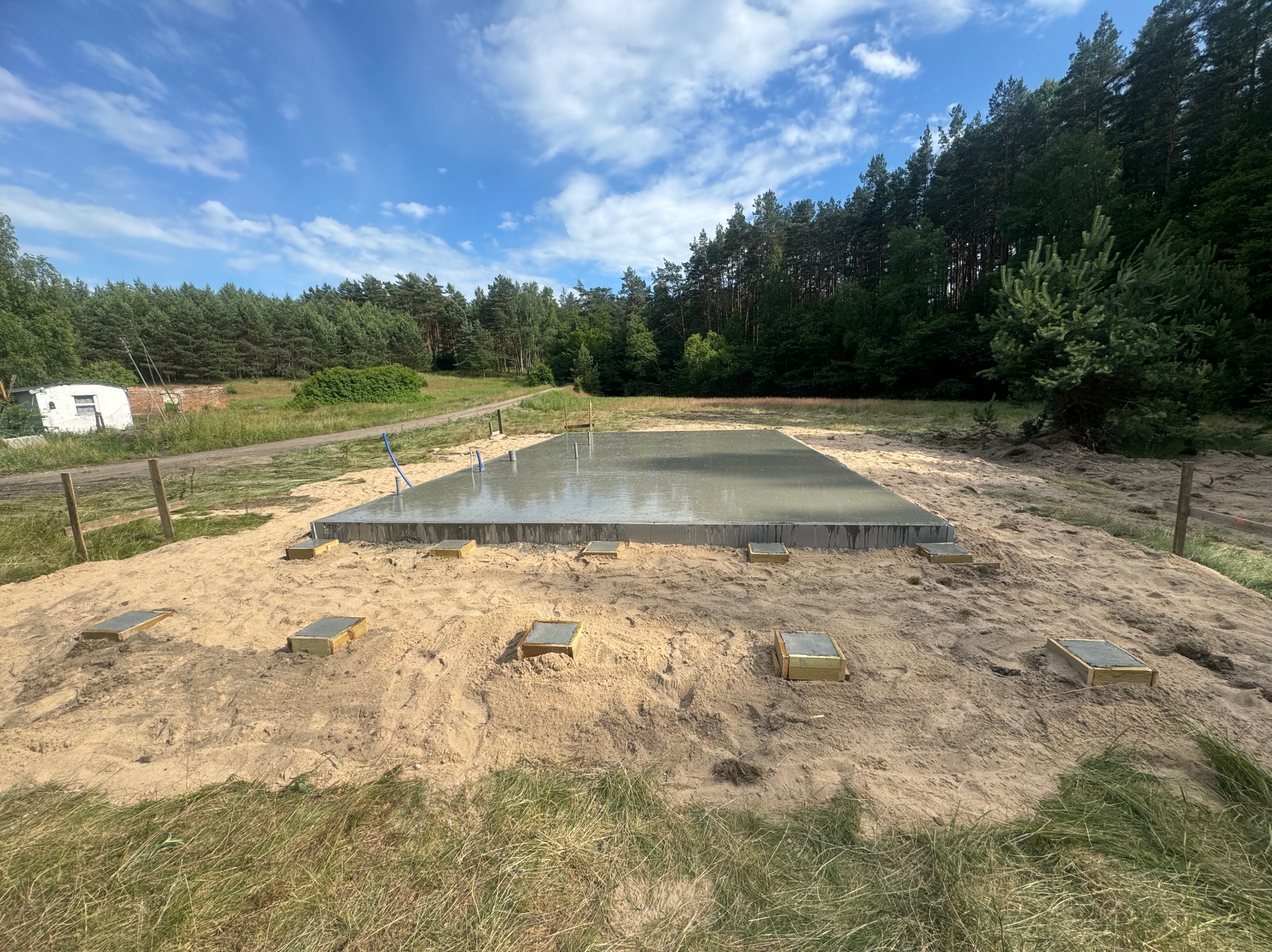 Świeżo wylana, mokra płyta fundamentowa na piaszczystym podłożu z drewnianymi podkładkami, widok z poziomu gruntu, w tle las i błękitne niebo z chmurami.