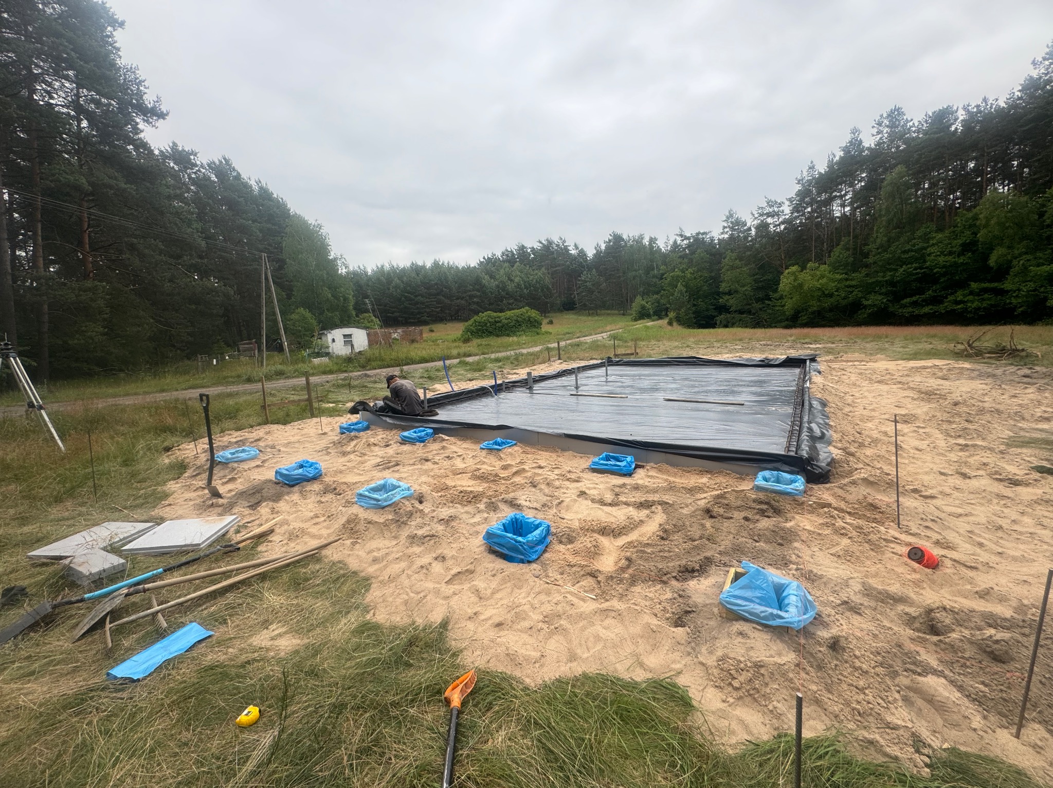 Przygotowanie terenu pod wylewkę fundamentów: rozłożona folia hydroizolacyjna, wyznaczone punkty, narzędzia i materiały budowlane na piaszczystym podłożu, widok na las w tle.