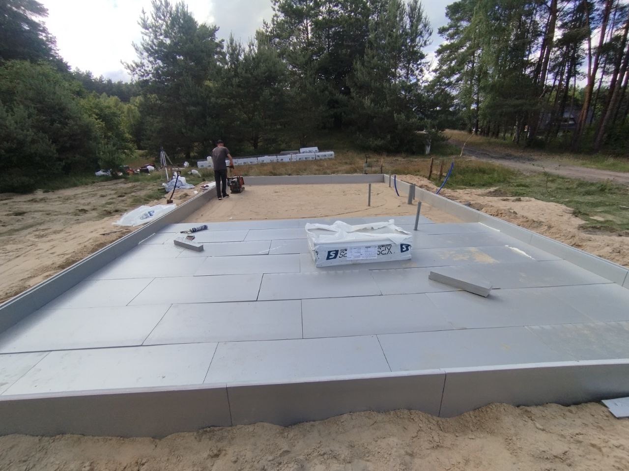 Przygotowany teren pod wylewkę fundamentów z ułożonymi płytami izolacyjnymi XPS, widoczny pracownik z zagęszczarką oraz elementy szalunku. W tle las i niebo.