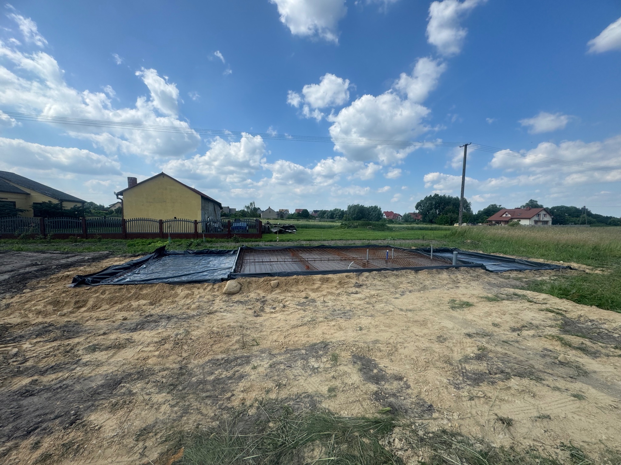 Przygotowanie zbrojenia pod płytę fundamentową w Warszawie, widoczne rury instalacyjne, czarna folia i wykop z piasku pod przyszły budynek mieszkalny w słoneczny dzień.