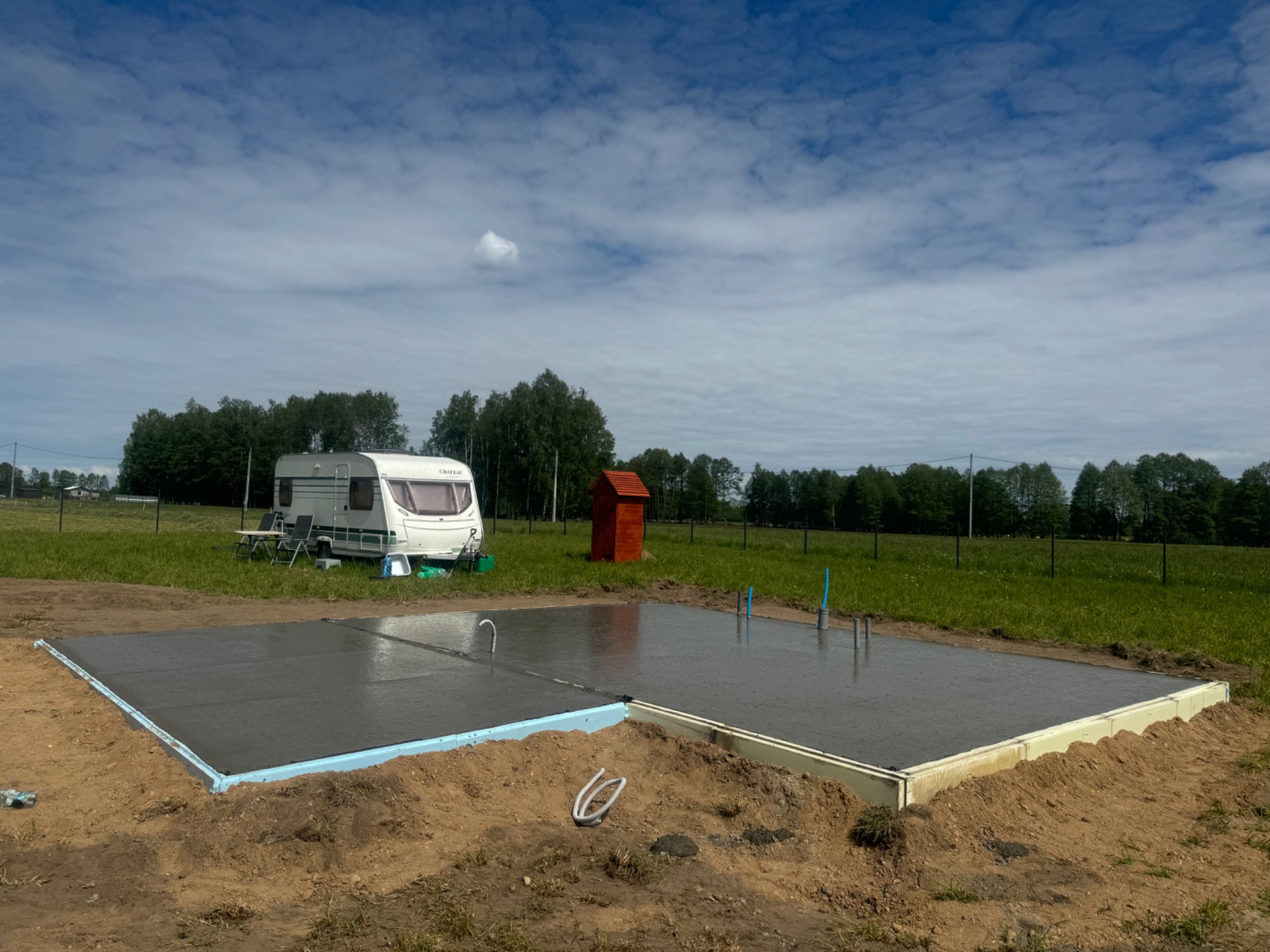 Świeżo wylany, gładki betonowy fundament pod budynek z widoczną izolacją termiczną krawędzi, rurami instalacyjnymi wystającymi z betonu i przyczepą kempingową w tle, na tle zielonej łąki...