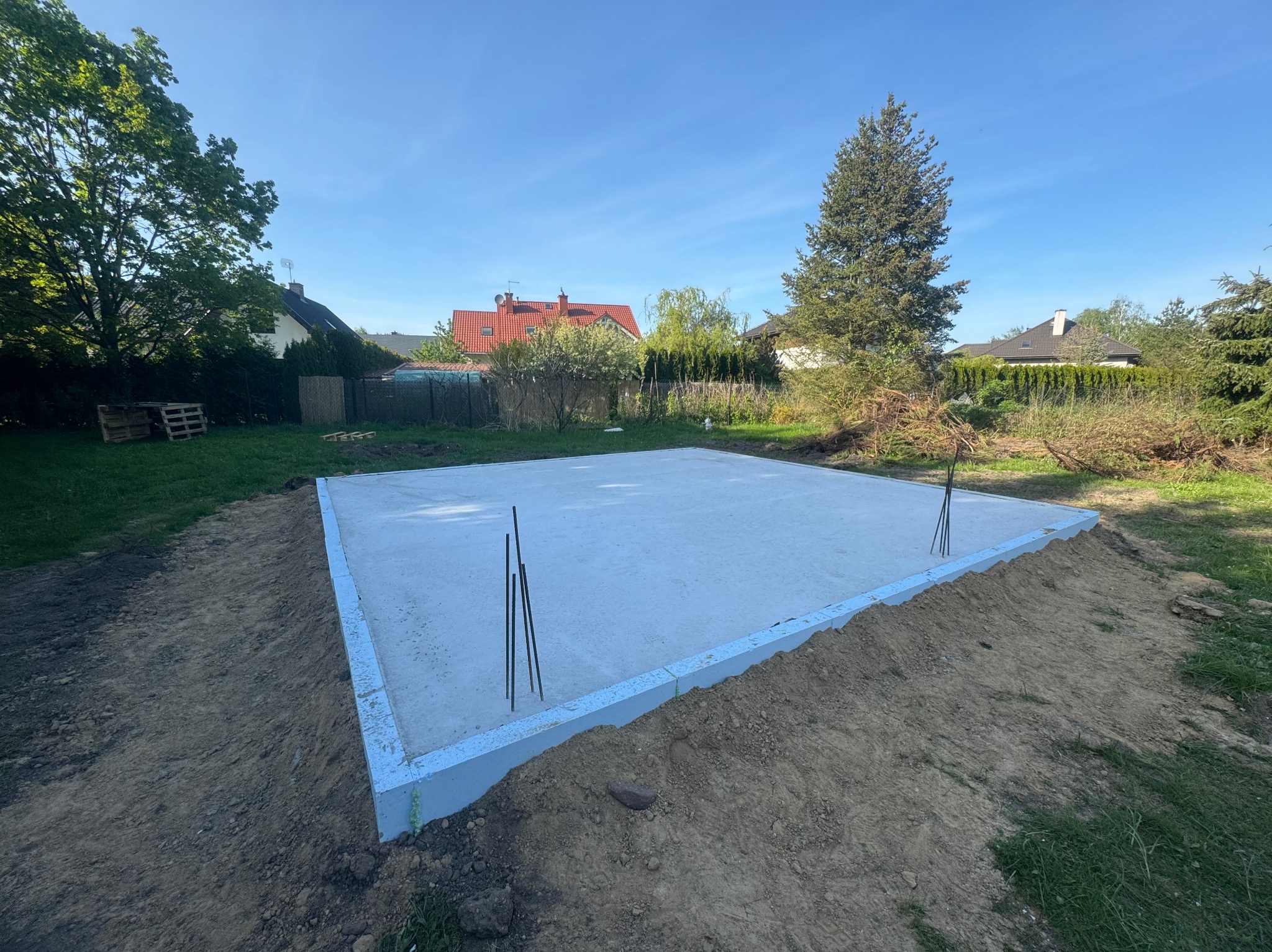 Wylany fundament płyty betonowej z izolacją obwodową w kolorze jasnoniebieskim, wystające pręty zbrojeniowe, w tle trawa, drzewa i domy pod błękitnym niebem.