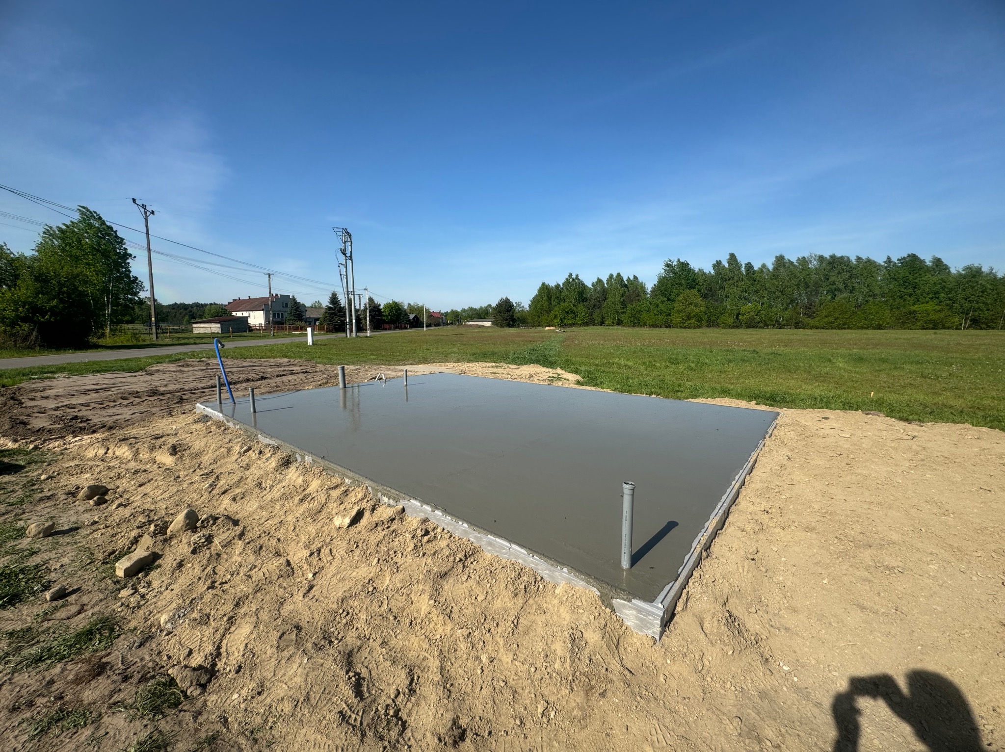Świeżo wylana, gładka płyta betonowa fundamentowa z widocznymi pionowymi rurami instalacyjnymi, otoczona piaskiem, na tle zielonego pola i błękitnego nieba.