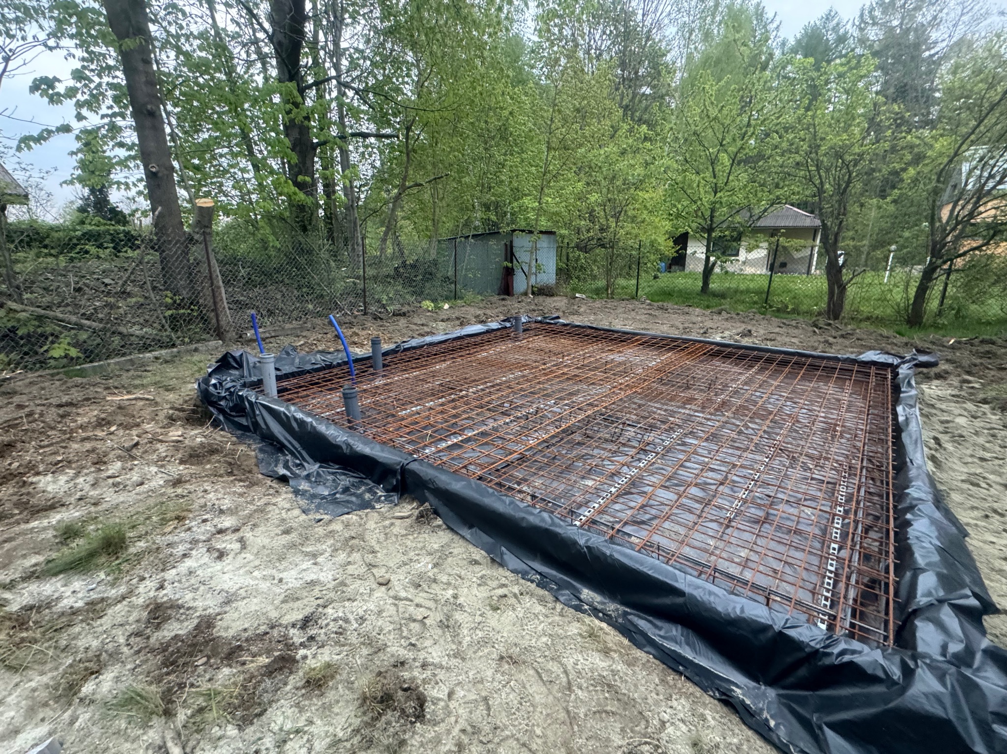 Przygotowany szkielet zbrojeniowy na czarnej folii pod wylewkę, widoczne wystające rury kanalizacyjne, w tle drzewa i ogrodzenie z siatki.