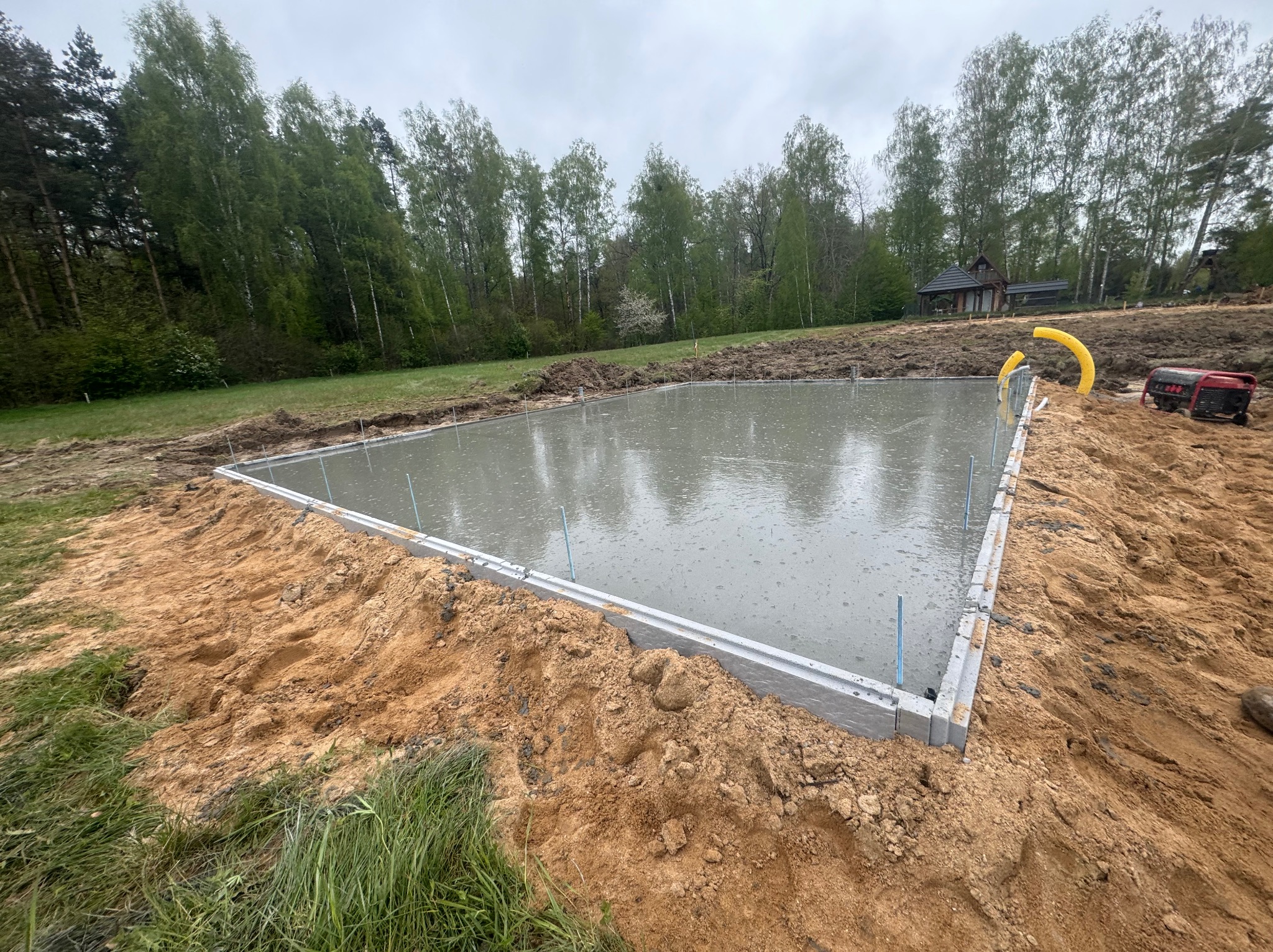 Wylany beton na przygotowany fundament, widoczne szalunki, rury i agregat prądotwórczy na tle zieleni i drzew