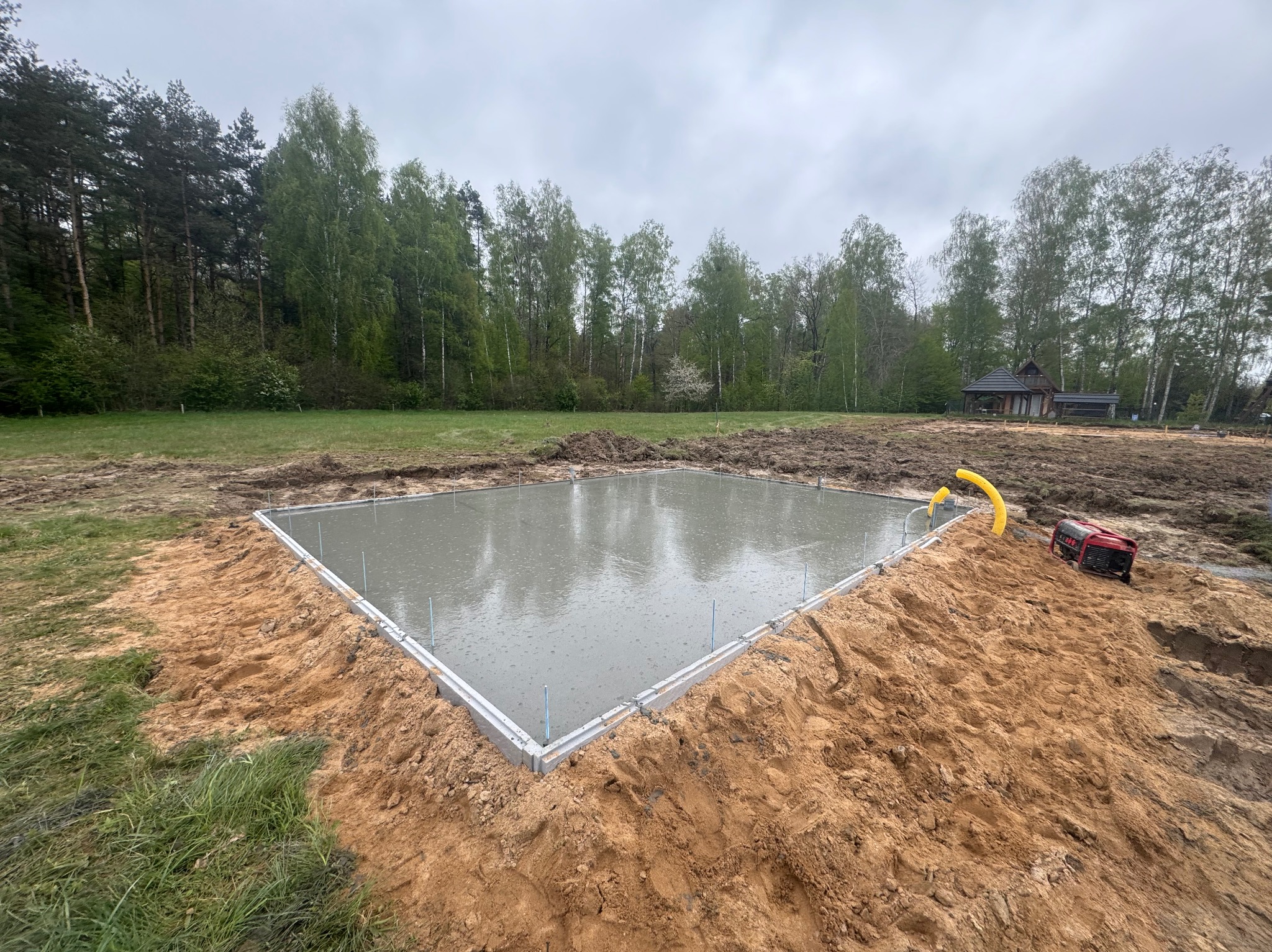 Świeżo wylana płyta betonowa z szalunkiem i wystającymi prętami zbrojeniowymi, otoczona piaskiem i trawą, z widocznym agregatem prądotwórczym i lasem w tle w pochmurny dzień.