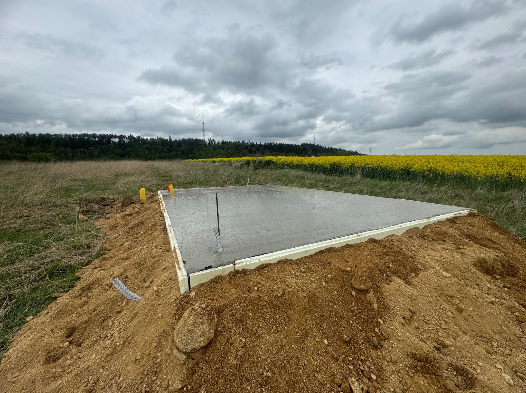 Świeżo wylana płyta betonowa pod budynek, otoczona ziemią i widokiem na pole rzepaku pod pochmurnym niebem.