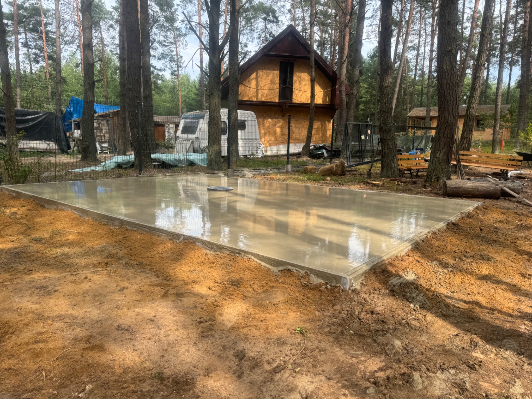 Świeżo wylany fundament pod budynek w lesie, widoczne odbicia drzew w mokrym betonie.