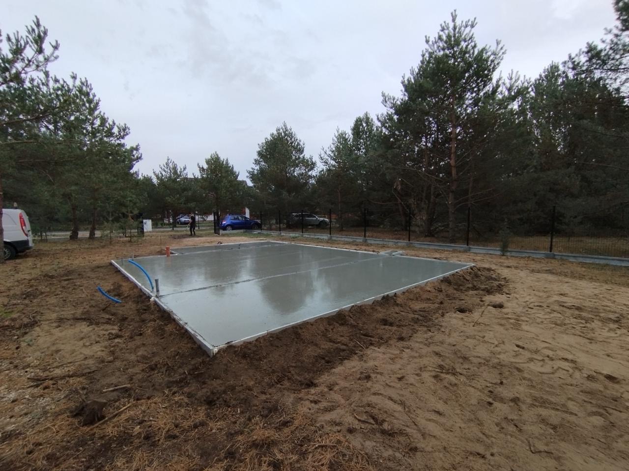 Wylany, świeży betonowy fundament pod budowę domu jednorodzinnego na piaszczystej działce z widocznymi drzewami iglastymi w tle.