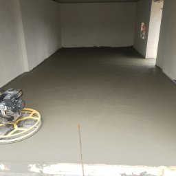 SEKO-Sebastian Kozioł - Zacieraczka mechaniczna na świeżo wylanej, szarej posadzce betonowej w przestronnym, nieukończonym pomieszczeniu z jasnymi ścianami, ukazująca etap prac wykończeniowych.