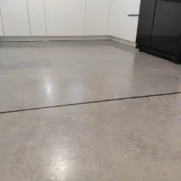 Szara, gładka posadzka w kuchni z białymi szafkami i czarną lodówką, widoczna dylatacja.