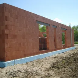 Wznoszona ściana domu z pustaków ceramicznych z widocznymi otworami okiennymi i stalowymi nadprożami, ocieplona fundamentowa w gruncie.
