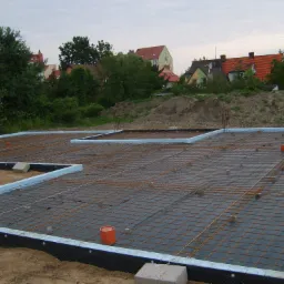 Przygotowany zbrojony fundament pod budynek, widoczna siatka zbrojeniowa, izolacja termiczna z polistyrenu ekstrudowanego i folia hydroizolacyjna, w tle zabudowa mieszkalna.