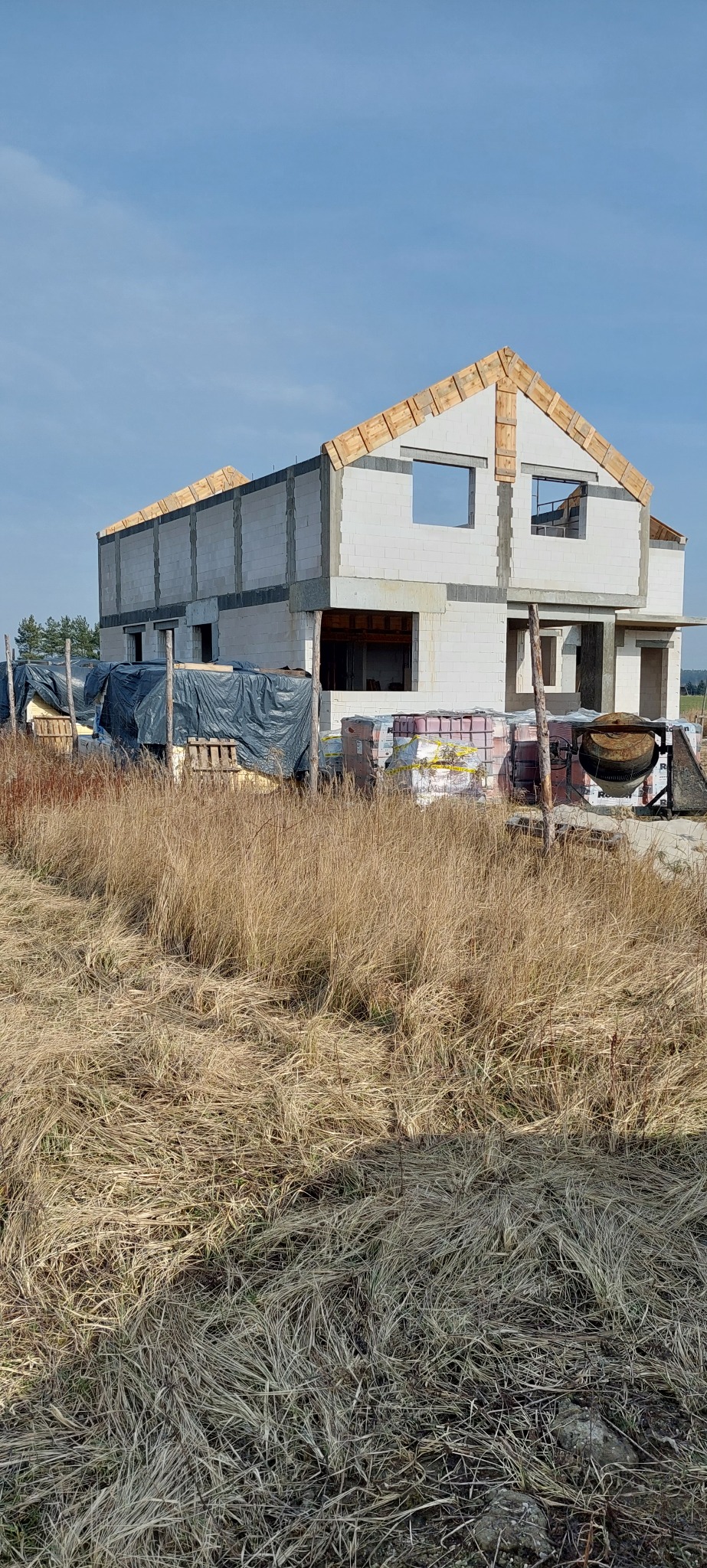 Niedokończony dwukondygnacyjny dom z białych bloczków, widoczne drewniane elementy konstrukcji dachu, na pierwszym planie wysoka trawa, w tle błękitne niebo.