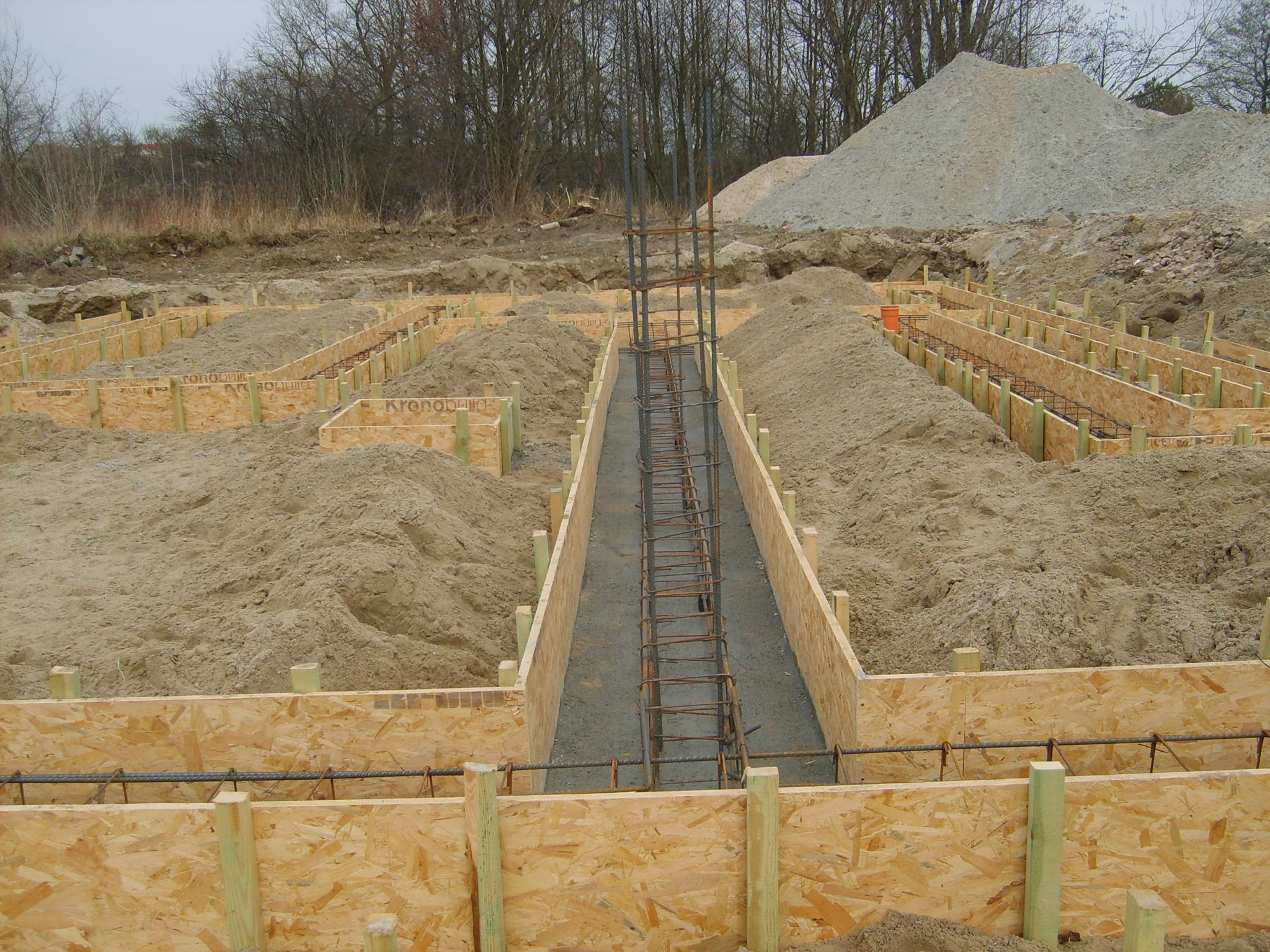 Szalunki z płyty OSB przygotowane pod wylewkę fundamentów, z widocznym zbrojeniem pionowym i poziomym, na nierównym terenie budowy.