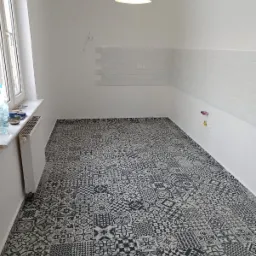 Wykończona kuchnia z wzorzystą, czarno-białą podłogą z płytek, białymi ścianami, częściowo wyłożonymi białymi płytkami typu metro, oraz czarną lampą wiszącą.