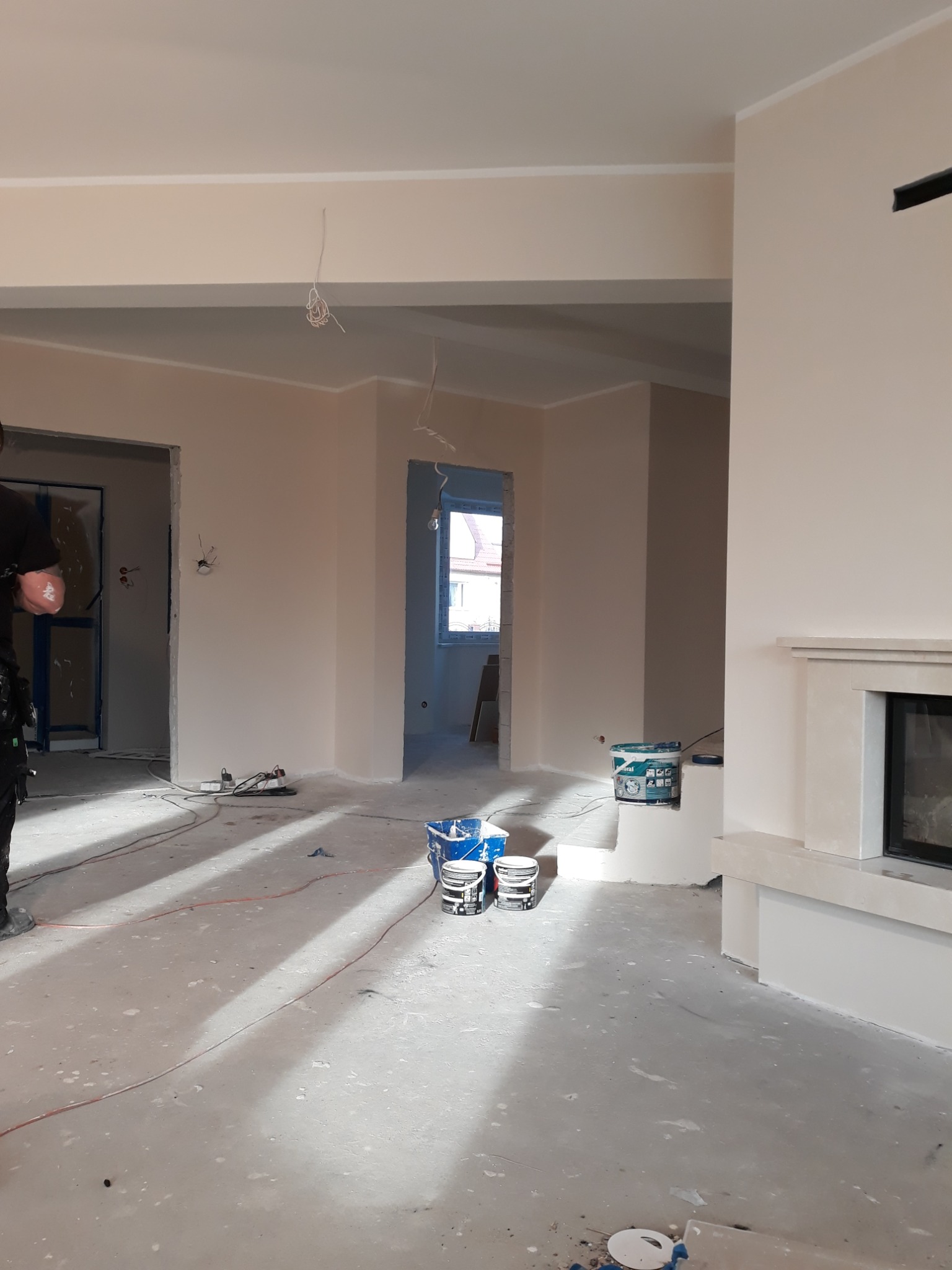 Pokój w trakcie remontu, ściany pomalowane na jasny kolor, widoczne puszki z farbą, kable wystające ze ścian i sufitu, przygotowany kominek, podłoga zabezpieczona folią.