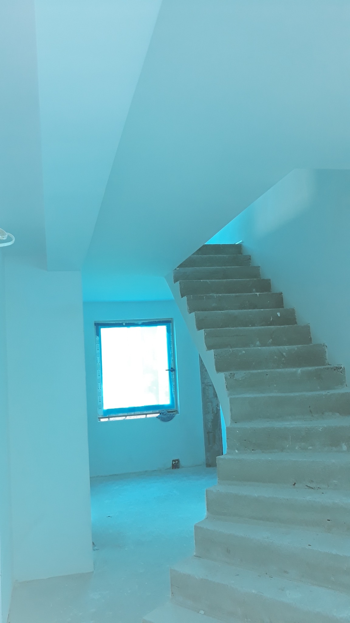 Wnętrze w trakcie prac wykończeniowych: surowe betonowe schody, okno zabezpieczone folią, ściany przygotowane do malowania.