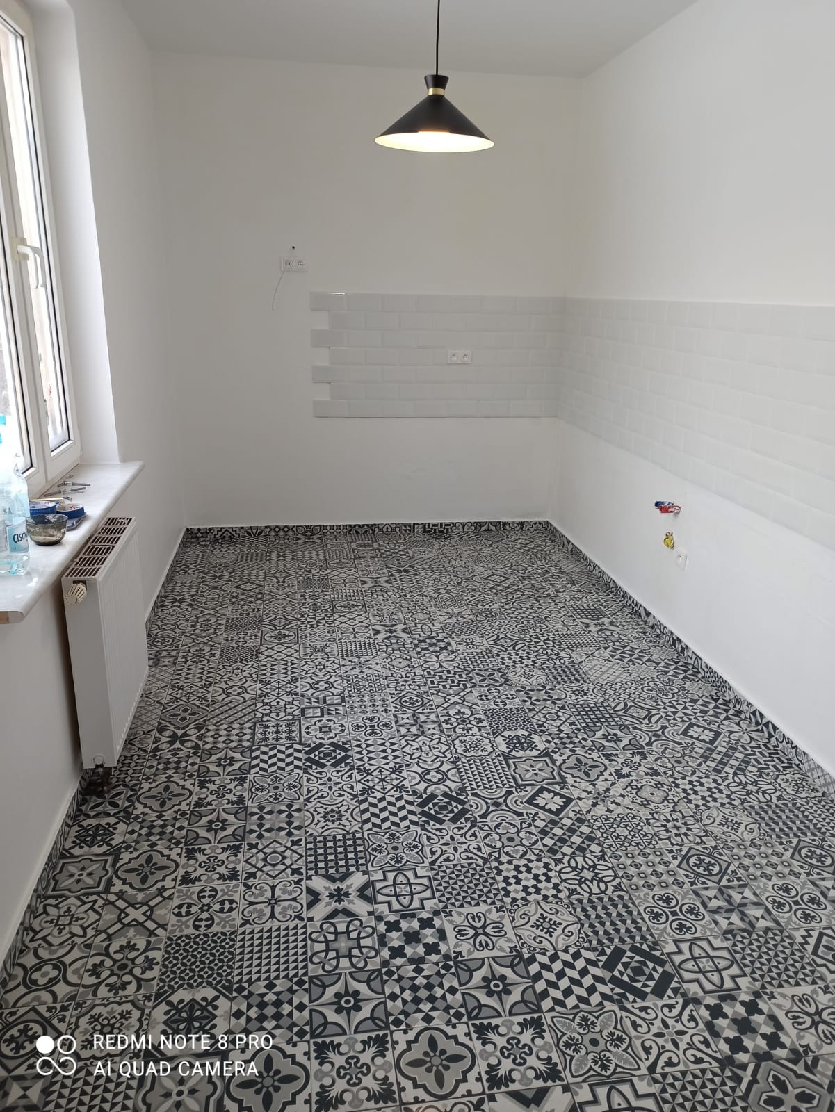 Wykończona kuchnia z wzorzystą, czarno-białą podłogą z płytek, białymi ścianami, częściowo wyłożonymi białymi płytkami typu metro, oraz czarną lampą wiszącą.