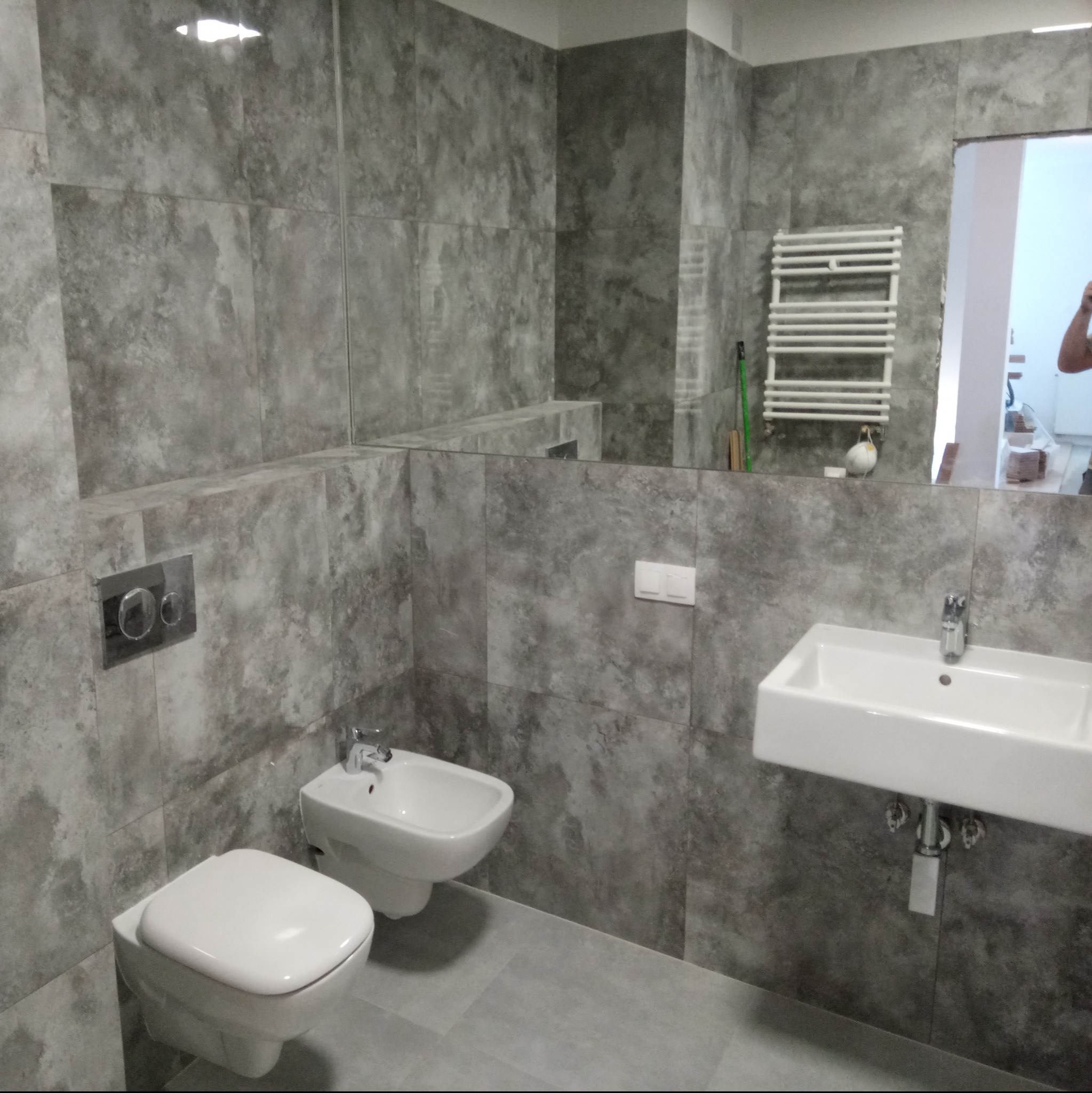 Wykończona łazienka z szarymi płytkami imitującymi beton, wisząca toaleta, bidet i umywalka, widoczne odbicie grzejnika drabinkowego w lustrze.