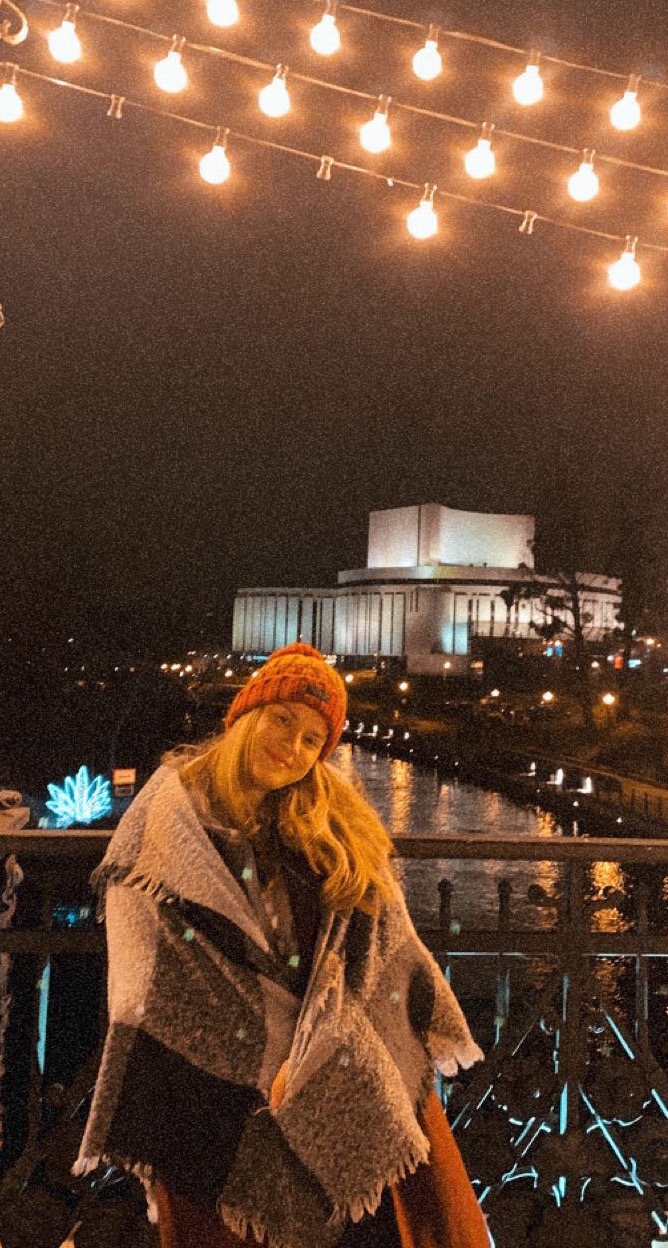 Uśmiechnięta młoda kobieta w pomarańczowej czapce i pledzie na tle oświetlonego budynku i rzeki nocą, dekoracyjne lampki nad głową.