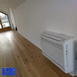 Biała jednostka wewnętrzna klimatyzacji podłogowej, model Hyper Inverter, zamontowana przy ścianie w jasnym pomieszczeniu z drewnianą podłogą i oknem dachowym w tle. W lewym dolnym rogu widoczne...