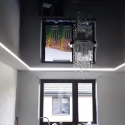 Nowoczesne wnętrze pokoju z błyszczącym, czarnym sufitem podwieszanym, oświetleniem LED na obwodzie, kryształowym żyrandolem i oknami z widokiem na zewnątrz.