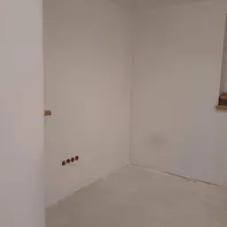 Pokój w trakcie przygotowania do malowania: zagruntowane ściany, widoczne puszki elektryczne i narzędzia malarskie na podłodze.