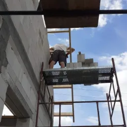 Murarz na rusztowaniu w trakcie wznoszenia ściany z białych bloczków, widok z dołu na tle błękitnego nieba.