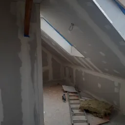 Poddasze w trakcie wykańczania: widoczne płyty gipsowo-kartonowe, wełna mineralna, drabina i okno dachowe.