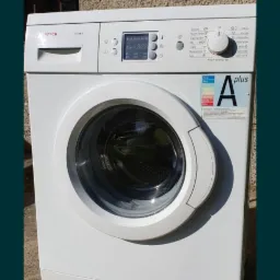 Biała pralka marki Bosch z widoczną klasą energetyczną A plus, ustawiona na zewnątrz, na tle szarej ściany z fakturą.