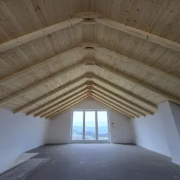 Poddasze z drewnianą konstrukcją dachu, widok na trzyczęściowe okno z widokiem na krajobraz, ściany w trakcie wykończenia, surowa podłoga.