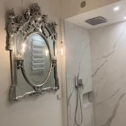 Nowoczesna łazienka z marmurowymi płytkami, deszczownicą i ozdobnym, srebrnym lustrem w stylu vintage, w którym odbija się grzejnik drabinkowy.