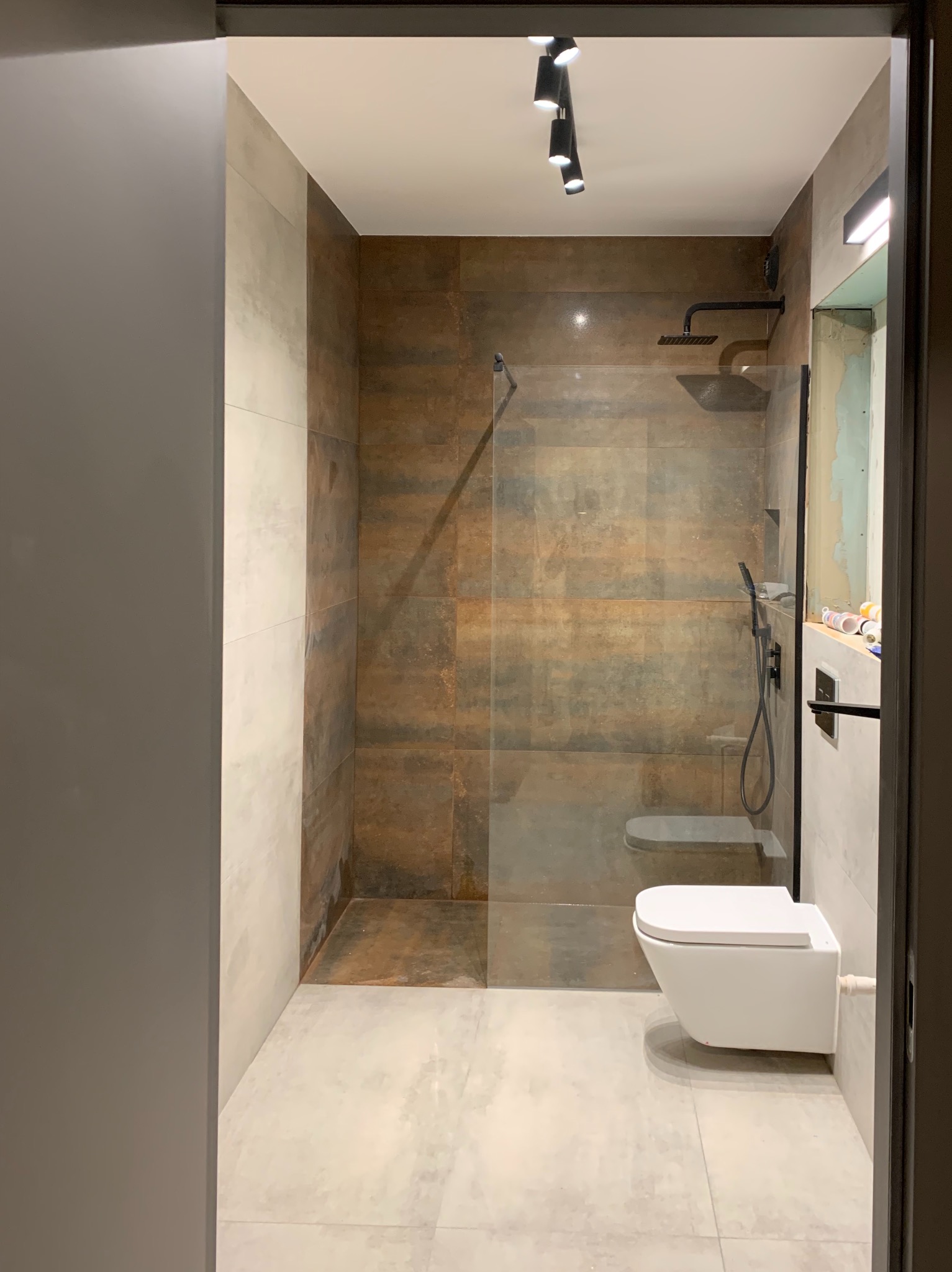 Nowoczesna łazienka z kabiną prysznicową typu walk-in, wykończona płytkami imitującymi rdzę, z podwieszaną toaletą i czarną armaturą, oświetlona reflektorami.