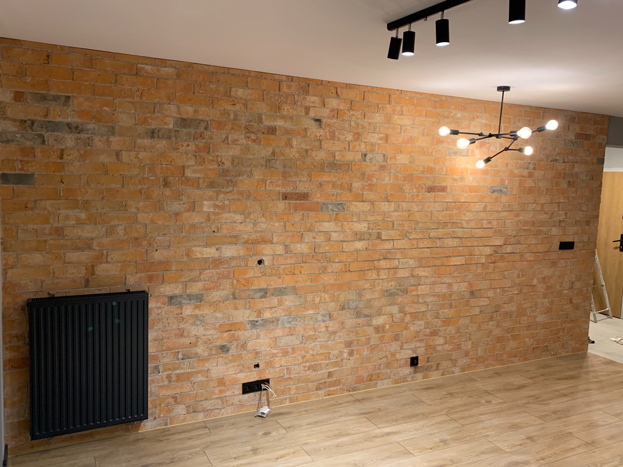 Pomarańczowa ściana z cegły w stylu loftowym, z widocznymi elementami instalacji elektrycznej w trakcie prac, czarny grzejnik, drewnopodobna podłoga i nowoczesne oświetlenie sufitowe.