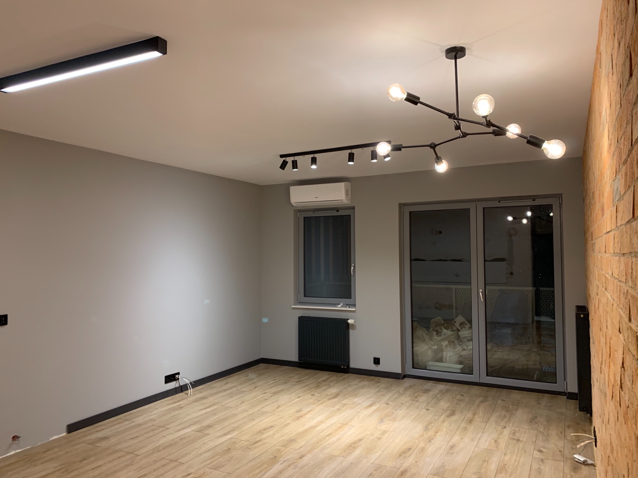 Wykończone wnętrze pokoju z parkietem, szarą ścianą, ceglaną ścianą, oknem, grzejnikiem i nowoczesnym oświetleniem sufitowym.