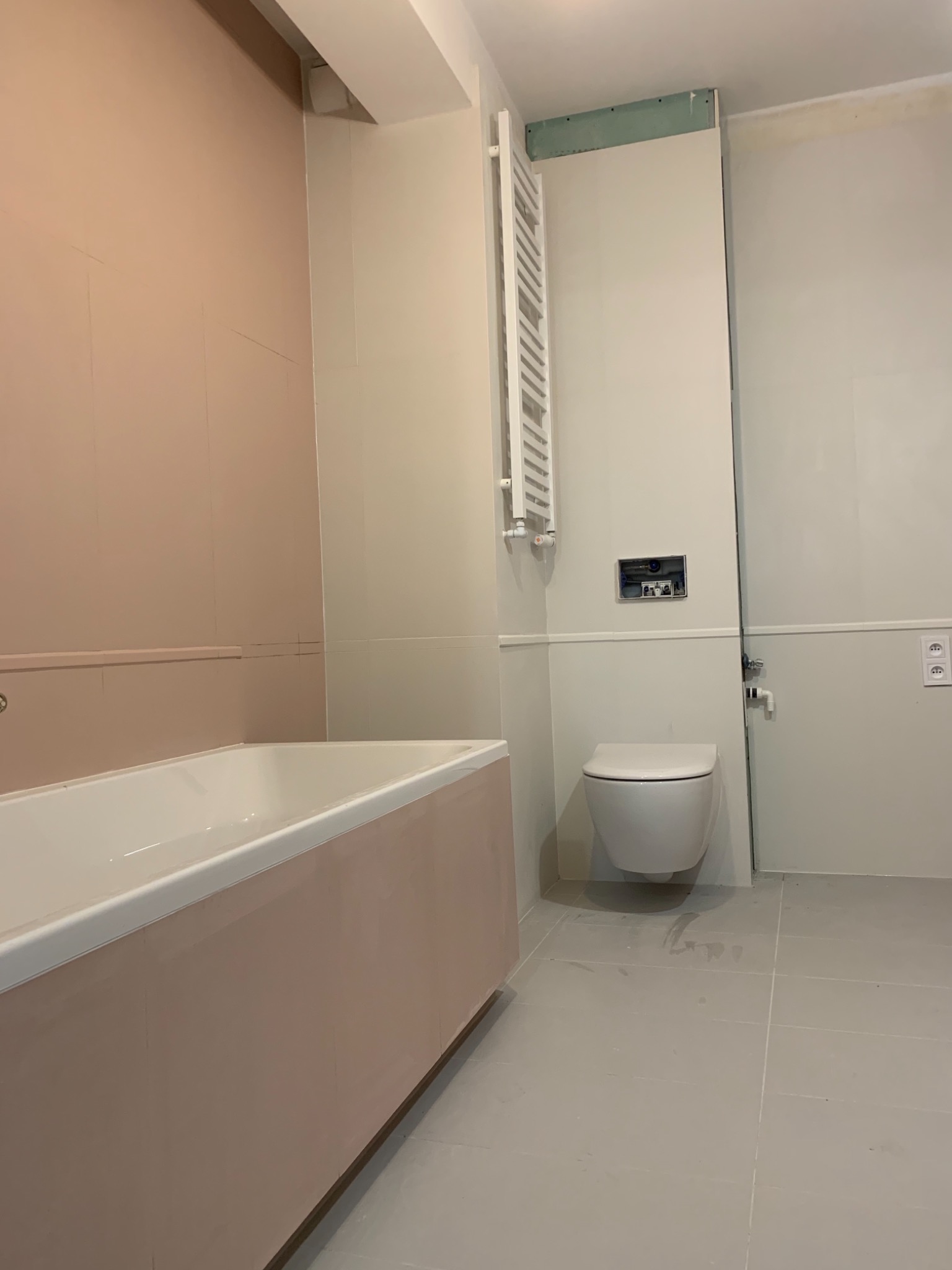 Remontowana łazienka z wanną w odcieniach różu, wiszącą toaletą, grzejnikiem drabinkowym i szarą podłogą z płytek, widoczne ślady po remoncie.