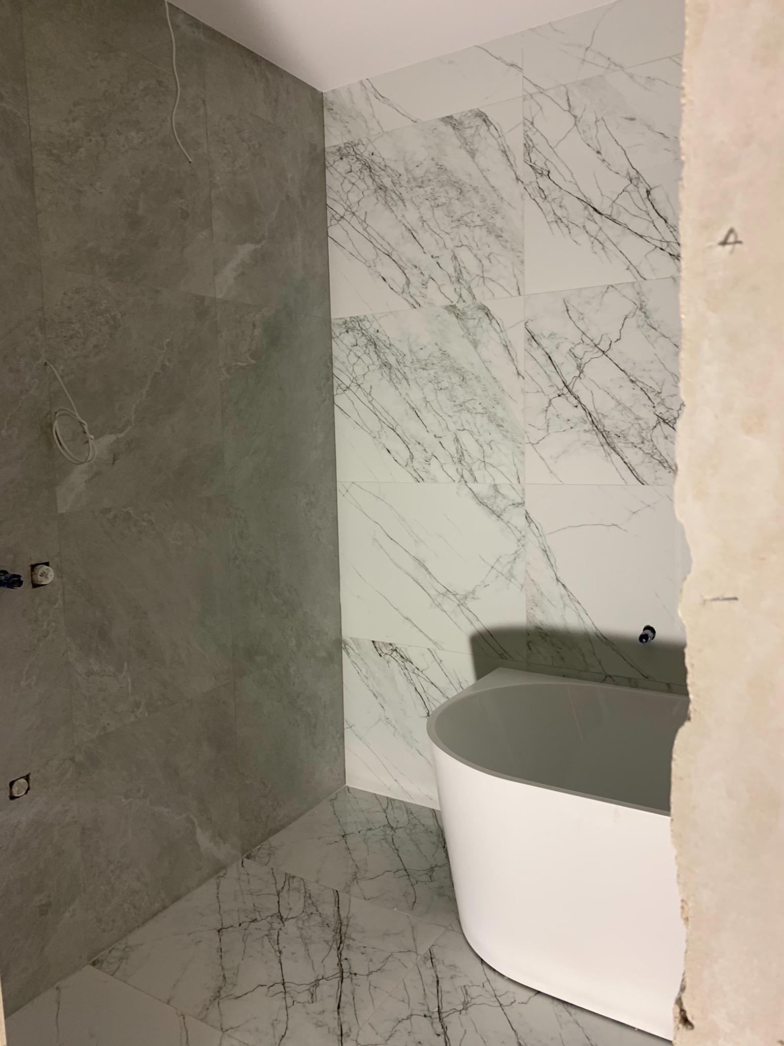 Łazienka w trakcie wykańczania w Krakowie, z częściowo położonymi płytkami imitującymi marmur na ścianach i podłodze, oraz białą, wolnostojącą wanną w rogu pomieszczenia, widoczne instalacje...