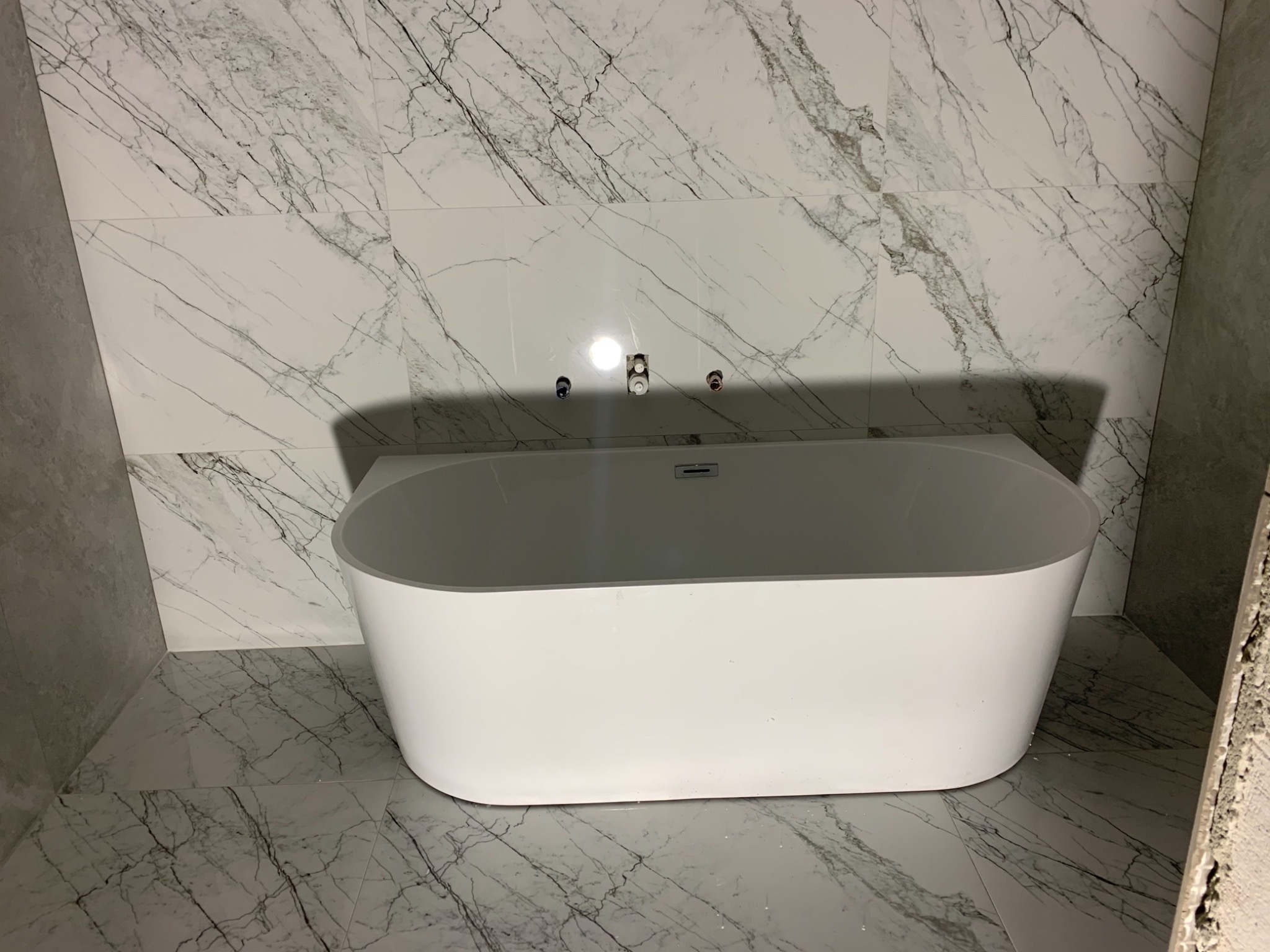 Wolnostojąca biała wanna na tle ściany i podłogi wyłożonych płytkami imitującymi biały marmur z szarymi żyłami, widoczne przygotowane przyłącza wodne.