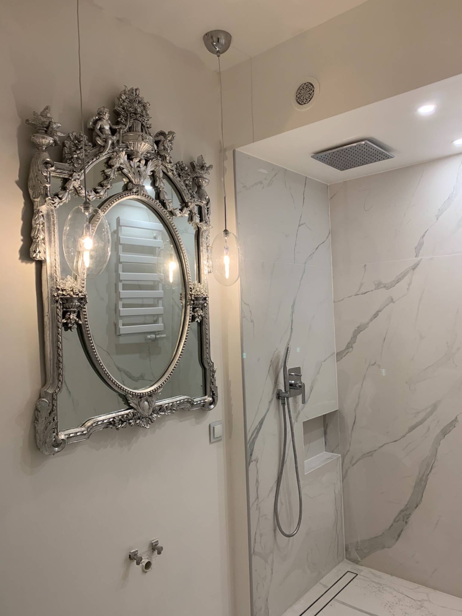Nowoczesna łazienka z marmurowymi płytkami, deszczownicą i ozdobnym, srebrnym lustrem w stylu vintage, w którym odbija się grzejnik drabinkowy.