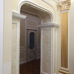 Eleganckie wnętrze: kolumna z pozłacanymi żłobieniami, łukowe przejście z dekoracyjną sztukaterią i drzwi w tle, ściany z beżową fakturą i dekoracyjnymi panelami.
