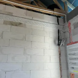 Rozprowadzenie przewodów elektrycznych w białych listwach na surowej ścianie z betonu komórkowego, widoczna skrzynka rozdzielcza z zabezpieczeniami, częściowo zamaskowana pianką montażową, na górze...