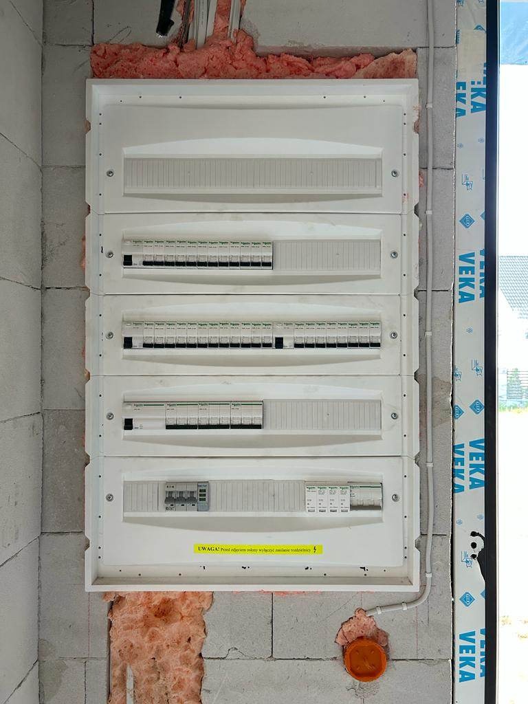 Rozdzielnica elektryczna zamontowana na surowej ścianie z betonu komórkowego, widoczne zabezpieczenia i okablowanie w trakcie montażu, z żółtą naklejką ostrzegawczą.
