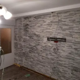 Ściana wewnętrzna wykończona szarą okładziną imitującą kamień, widoczne niedokończone prace elektryczne, kable wystające ze ściany, fragment podłogi z narzędziami i wiadro.