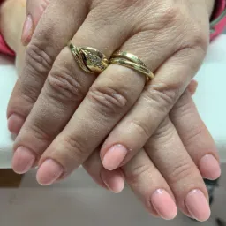 Dłonie kobiety ze świeżo wykonanym manicure żelowym w odcieniu pudrowego różu, zdobione złotymi pierścionkami.