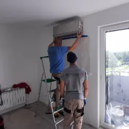 Dw&oacute;ch instalator&oacute;w montuje klimatyzator na ścianie w jasnym pomieszczeniu, jeden na drabinie, drugi obserwuje, widoczne narzędzia i zabezpieczona ściana.