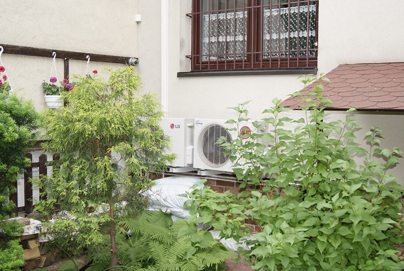Dwie jednostki zewnętrzne klimatyzacji LG Inverter zamontowane na ceglanym fundamencie, widoczne zza bujnej zieleni ogrodu przydomowego.