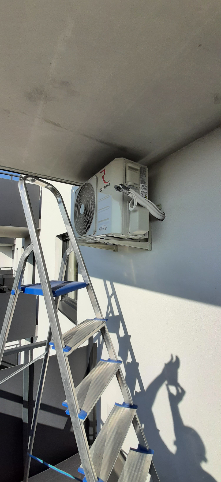 Zewnętrzna jednostka klimatyzacji Rotenso zamontowana na białej ścianie budynku, widoczna drabina aluminiowa z niebieskimi elementami i cień osoby pracującej przy instalacji.
