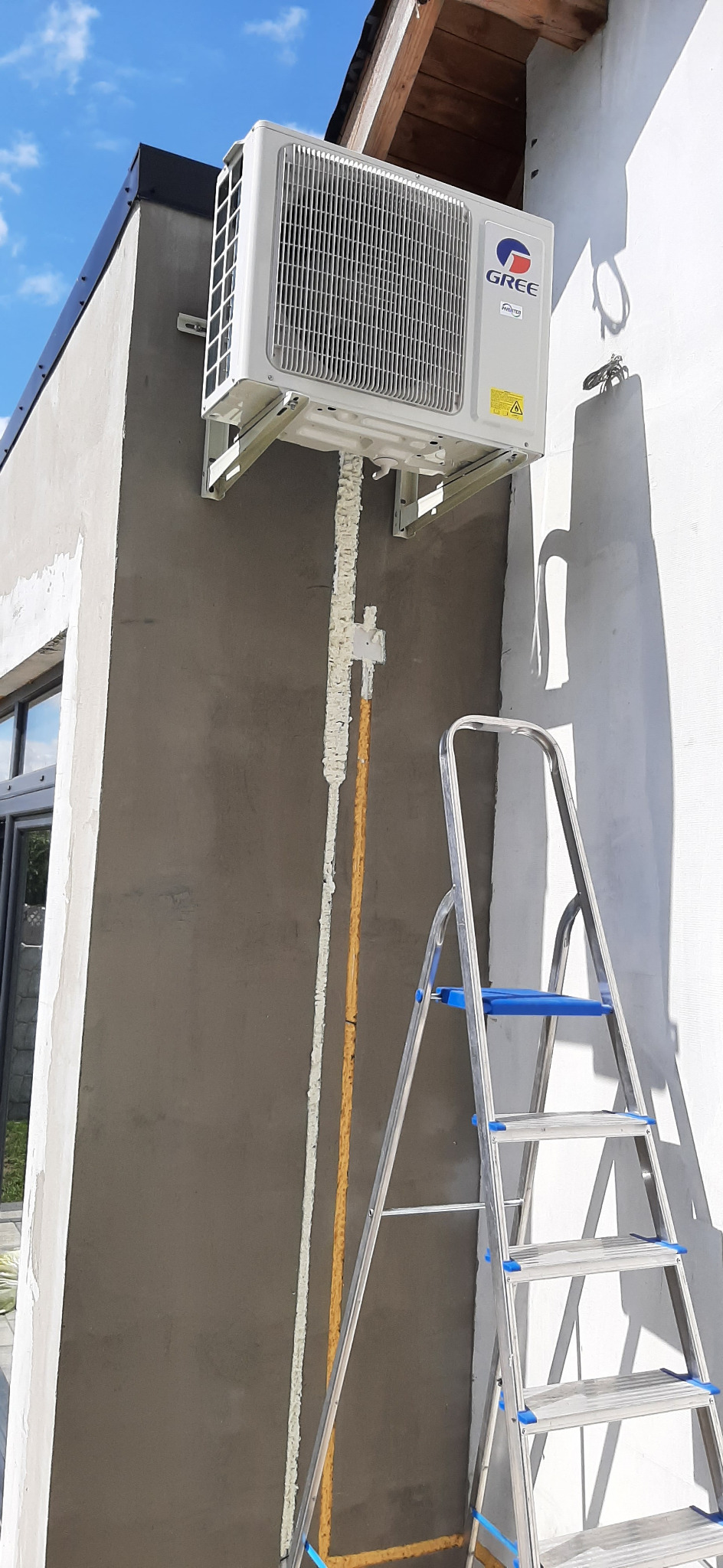 Zewnętrzna jednostka klimatyzacji Gree zamontowana na szarej ścianie budynku, rury zabezpieczone pianką montażową, drabina aluminiowa z niebieskimi elementami oparta o ścianę.